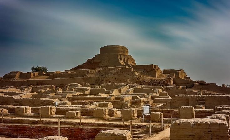 Wide-angle view of the excavated brick structures at Mohenjo-Daro featuring the famous central Buddhist stupa under a blue sky.