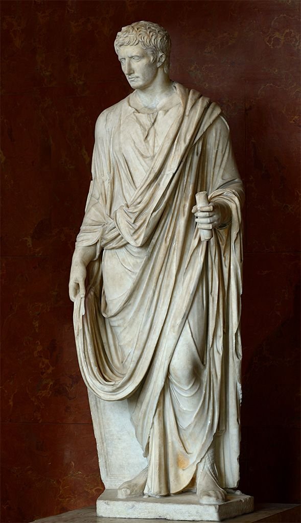 Full-length marble statue of Roman Emperor Augustus dressed in a flowing toga, holding a rolled scroll in his left hand against a dark reddish background.