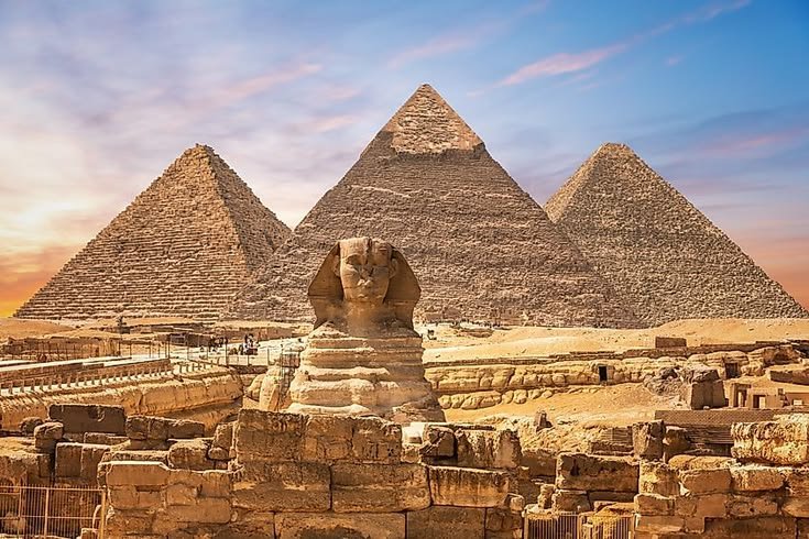 Front view of the Great Sphinx sitting in front of the three Pyramids of Giza in Egypt against a warm sunset sky with soft clouds.