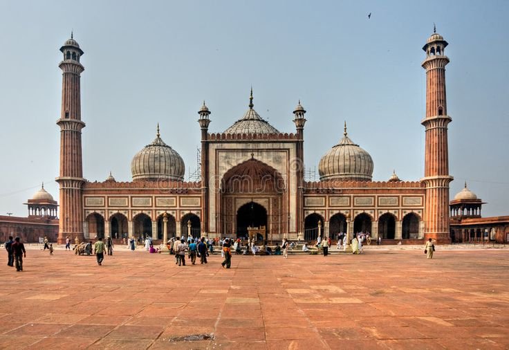 Wide shot of the red sandstone Jama Masjid with white marble domes, tall minarets, and visitors across the large open courtyard.