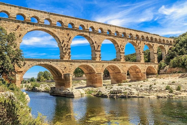 A photo of the ancient three-tiered Roman aqueduct Pont du Gard in Vers-Pont-du-Gard, France, spanning the Gardon River under a blue sky with white clouds. Green foliage frames the left side.