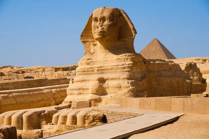 Full side profile of the limestone Great Sphinx of Giza sitting in the desert sand with a Great Pyramid rising in the background under a clear blue sky.