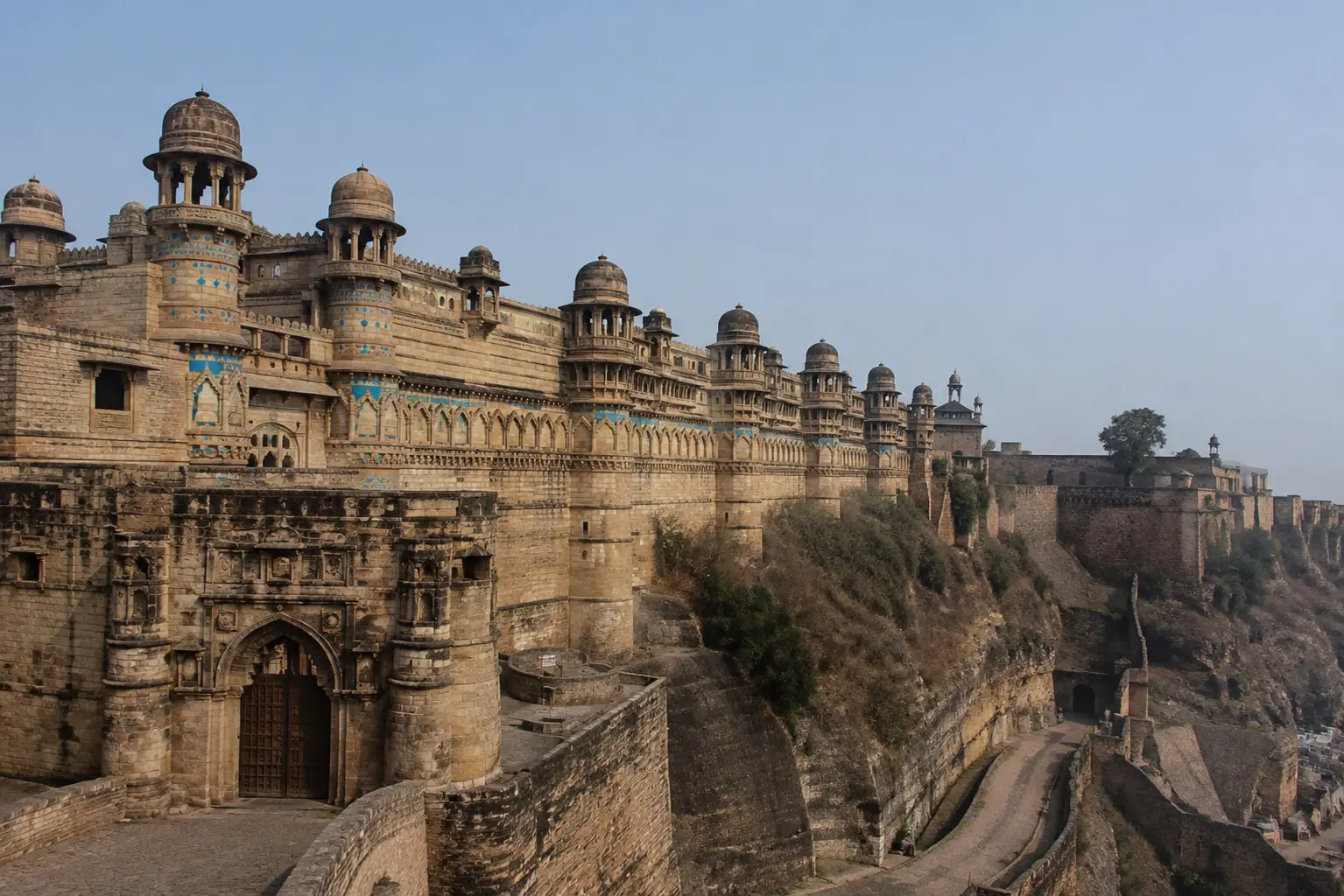 Stone outer walls and domed towers of Gwalior Fort decorated with blue ceramic tile work along a hilltop cliff.